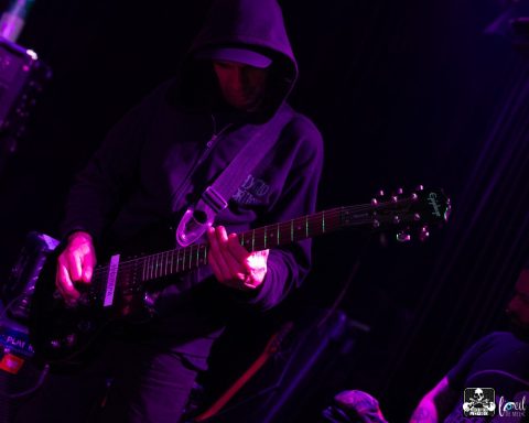 A musician in a black hoodie playing an electric guitar onstage.