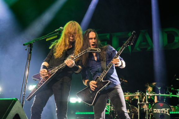 Two guitarists perform passionately on stage with bright lights behind them.