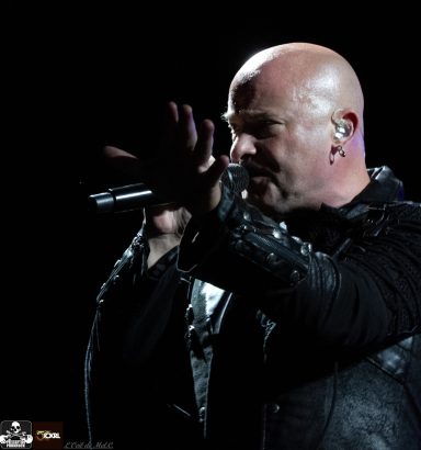 Bald singer performing with a microphone, wearing a black jacket against a dark background.