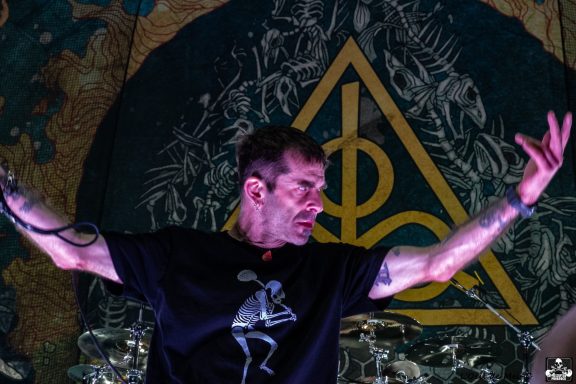 A performer on stage with arms raised, against a vibrant, decorative background.