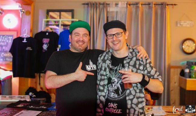 Two men posing with smiles, one wearing a black shirt and the other in a patterned jacket.