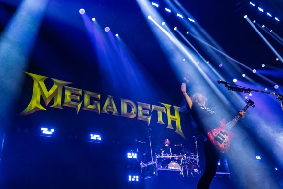A guitarist performing on stage with a large "Megadeth" logo in the background.