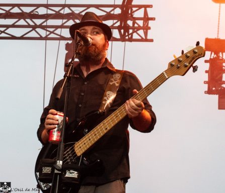 Musician with a beard playing bass guitar and holding a can at a concert.