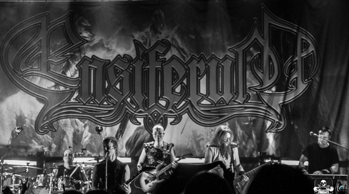 A black and white photo of a band performing on stage with a prominent logo backdrop.