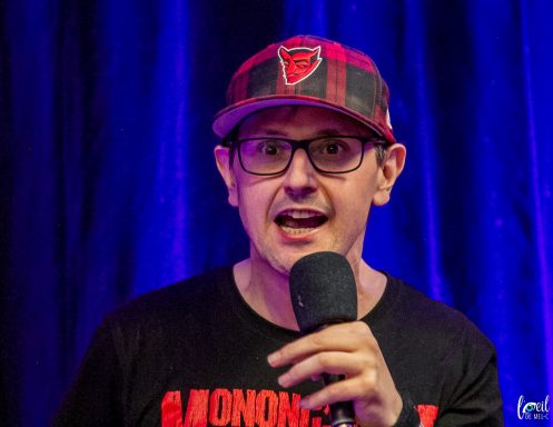 Man speaking into a microphone, wearing a plaid cap and a black t-shirt, against a blue backdrop.