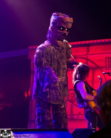 A performer in a large monster costume singing on stage with a guitarist nearby.
