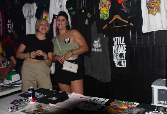 Two young women smile behind a table of T-shirts and merchandise in a dark setting.