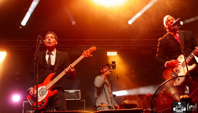 Two musicians perform on stage with instruments; a camera operator captures the moment.