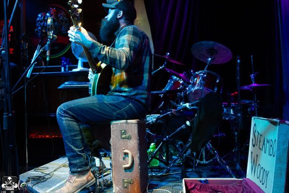 Musician playing guitar on stage, with drums visible in the background.
