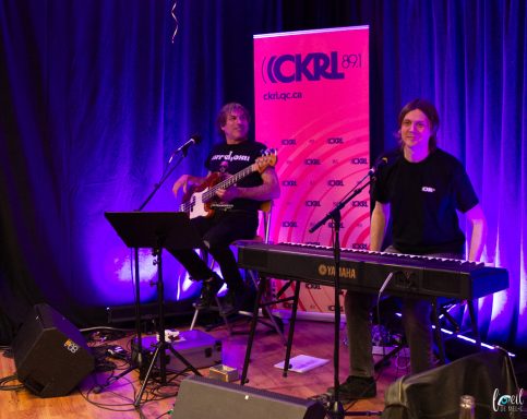 Two musicians performing live, one on guitar and one on keyboard, with a backdrop banner.