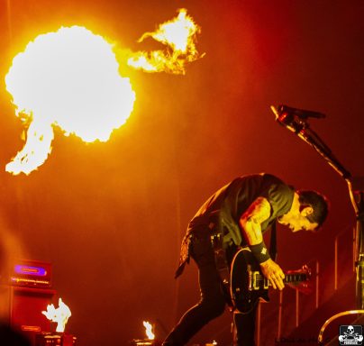 Musician playing guitar on stage with flames and dramatic lighting in the background.