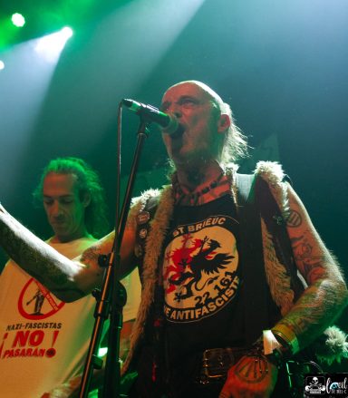 A musician with tattoos performs on stage, wearing a tank top with a bold graphic and holding a microphone.