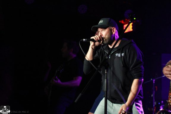 A vocalist performing on stage with a microphone, wearing a black cap and hoodie.