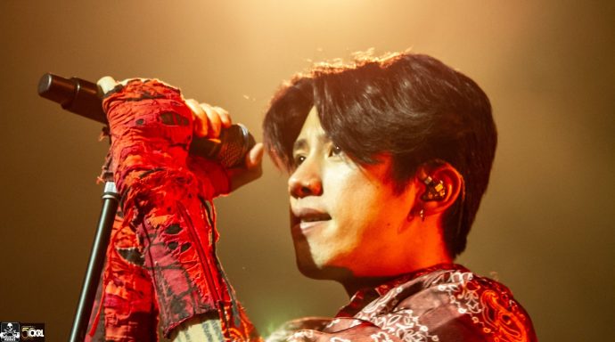 A singer with dark hair holds a microphone, wearing a red outfit, under dramatic lighting.
