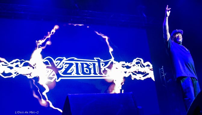 A performer on stage with a raised arm in front of a vibrant graphic backdrop.