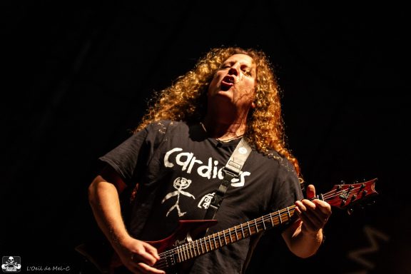 Musician playing electric guitar with long, curly hair, wearing a black T-shirt.