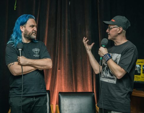 Rouge Pompier - Radiothon CKRL 89,1 Two men discussing on stage, one with blue hair and the other wearing glasses and a cap.