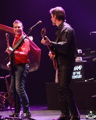 Two musicians performing on stage with instruments, illuminated by colorful stage lights.
