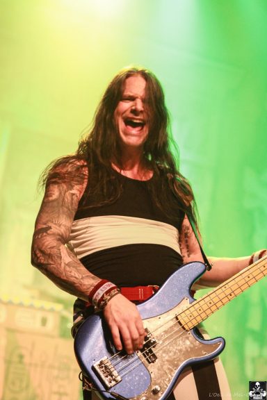 Musician with long hair plays a blue bass guitar, smiling on stage under green lights.