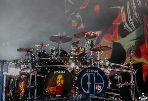 Drum set with artist logos, set against a dramatic smoky backdrop.
