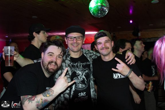 Three friends pose together at a lively party, holding drinks and making rock hand gestures.