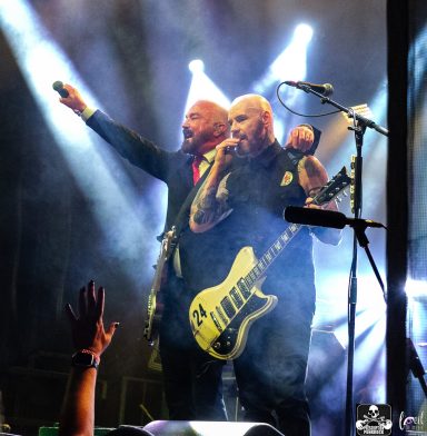Two musicians performing on stage with electric guitars and dramatic lighting.