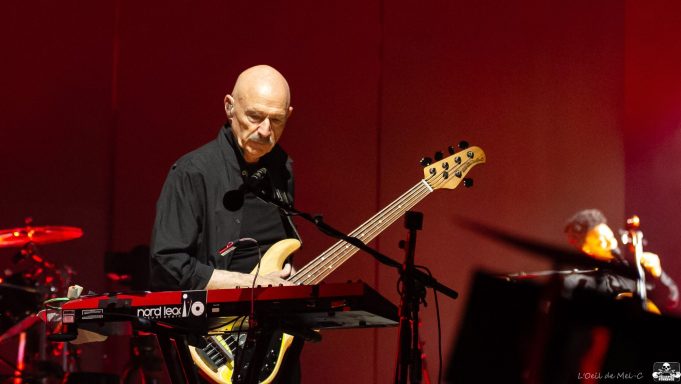 Musician playing an electric bass guitar on stage with instruments in the background.