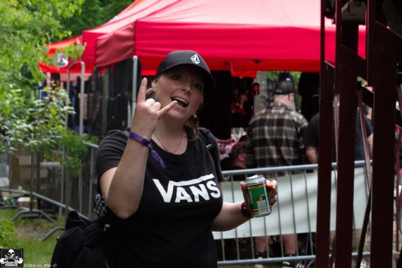 Person making a rock sign with one hand, holding a drink, in front of a festival tent.