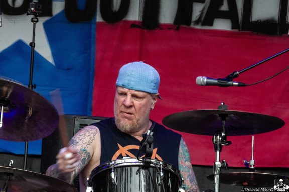 Drummer performing on stage, wearing a blue headband and surrounded by musical equipment.