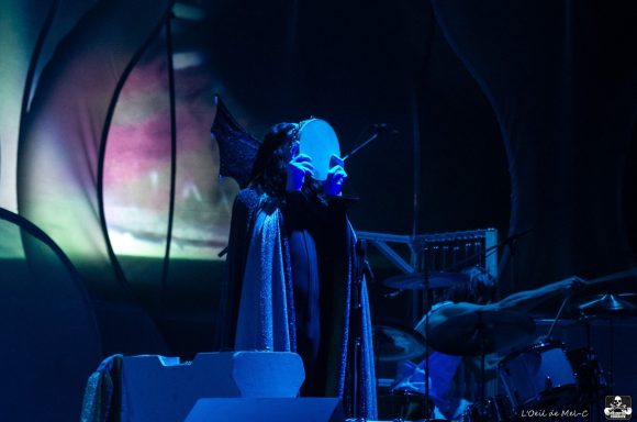 A performer in a dark costume with blue lighting on stage, surrounded by abstract shapes.
