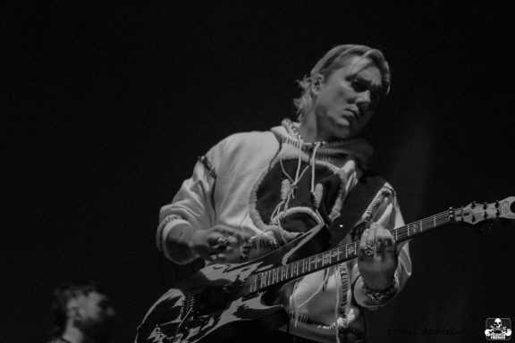 Musician playing electric guitar on stage, wearing a hoodie and focused on performance.