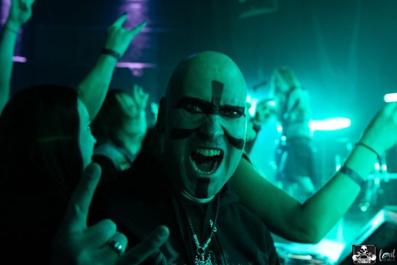 A man with face paint and a joyful expression holds up a rock hand gesture in a concert crowd.