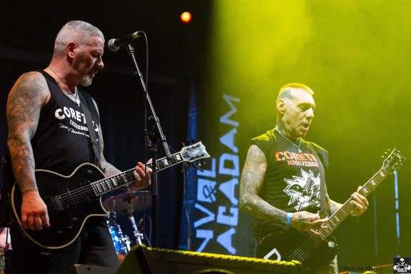 Two tattooed musicians perform on stage with electric guitars and dramatic lighting.