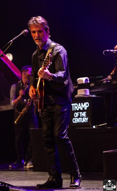 Musician performing on stage with a guitar, illuminated by purple lights.