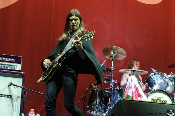 Musician playing guitar on stage with a drummer visible in the background.
