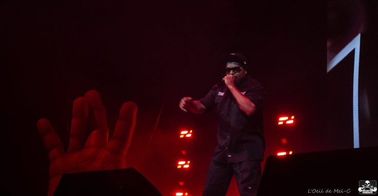 Performer on stage rapping, with dramatic lighting and a large hand sculpture in the background.