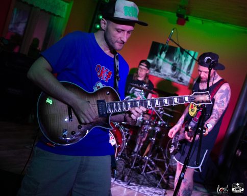 Musicians performing on stage, with a guitarist and drummers in a vibrant setting.