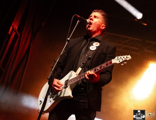 A singer performing on stage, holding a guitar with dramatic lighting in the background.