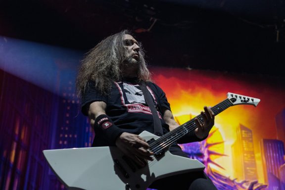 Musician playing electric guitar on stage with dramatic lighting in the background.