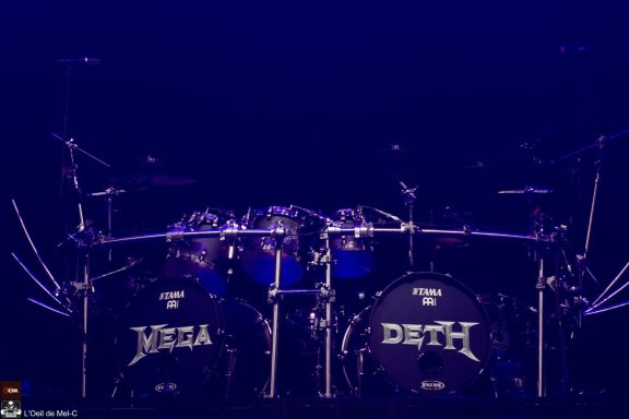 A drum set featuring large bass drums labeled "MEGA" and "DETH" against a dark backdrop.