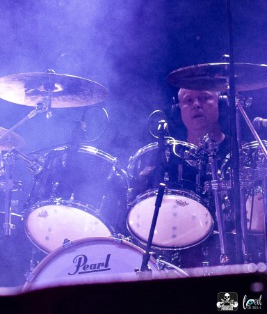 Drummer performing on stage, surrounded by fog and vibrant lighting.
