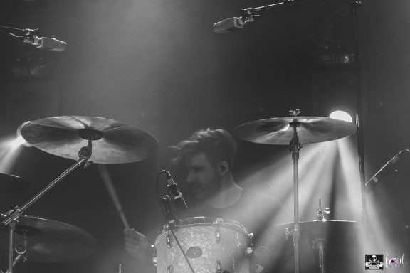 Drummer performing on stage with dramatic lighting effects in a monochrome setting.
