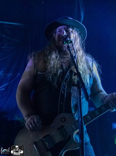 Musician with long hair wearing a hat, singing into a microphone with a guitar.