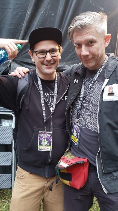 Two smiling men pose together, one pointing to his head, wearing event badges.
