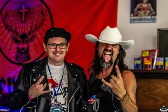 Two men pose with hand gestures, one wearing a white cowboy hat and the other in a leather jacket.