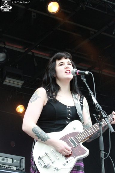 Woman with dark hair sings into a microphone while playing a white guitar on stage.
