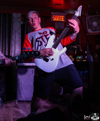 Musician performing energetically on stage with a white guitar.