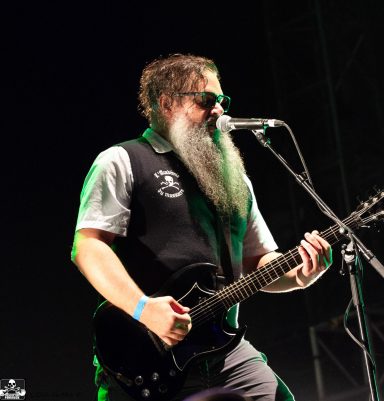 A musician with a beard playing electric guitar and singing on stage.