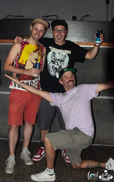 Three men posing playfully at an event, one kneeling and two standing, smiling.