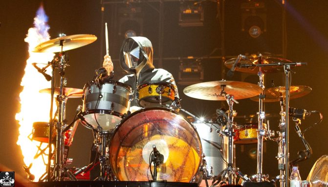 A drummer performing on stage with flames in the background.